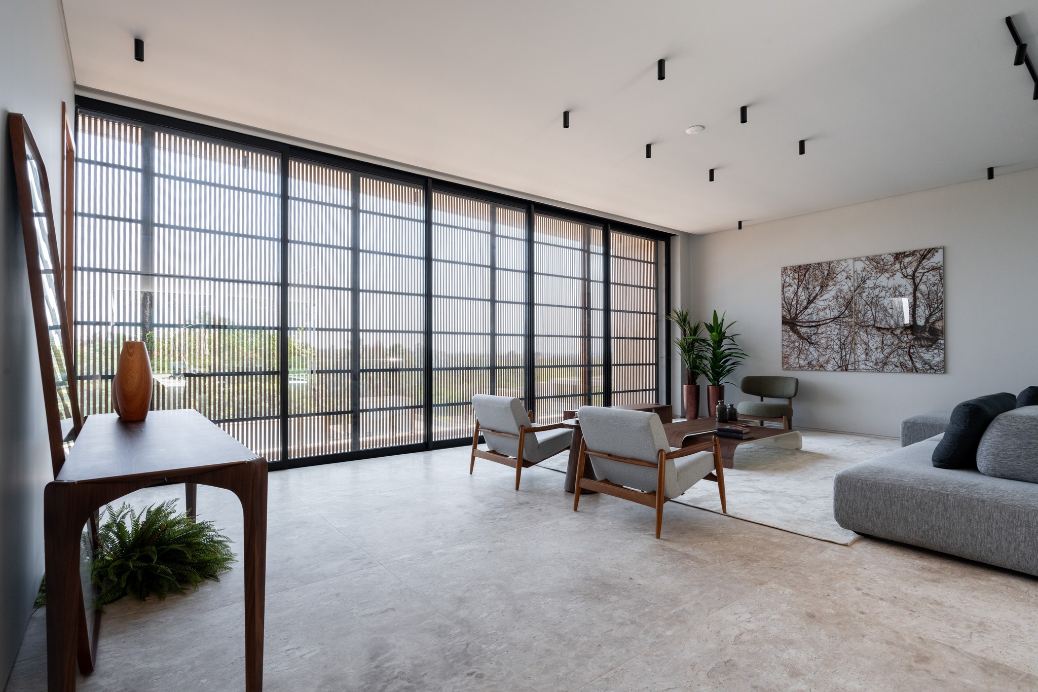 Sala de estar com poltronas de madeira, esquadrias em brise metálico e quadro fotográfico de galhos
