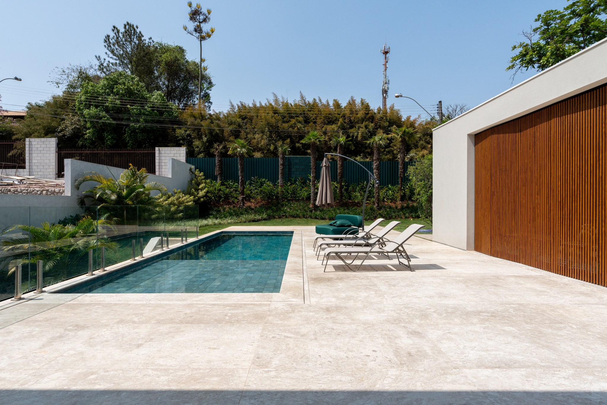 Piscina retangular com deque em travertino, espreguiçadeiras e vegetação tropical ao redor