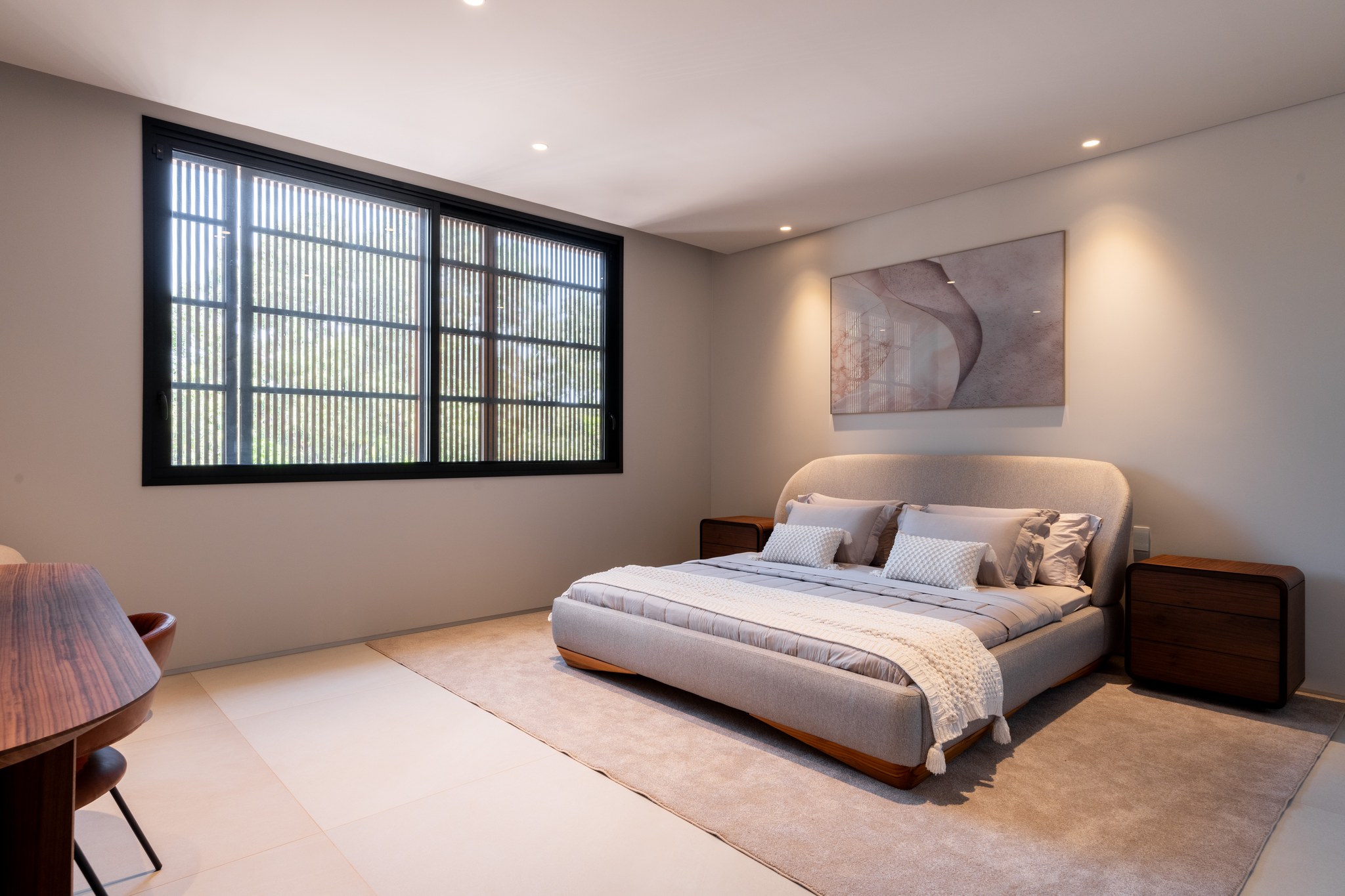 Suíte com cama de casal em tecido cinza, quadro abstrato e janela com brise metálico