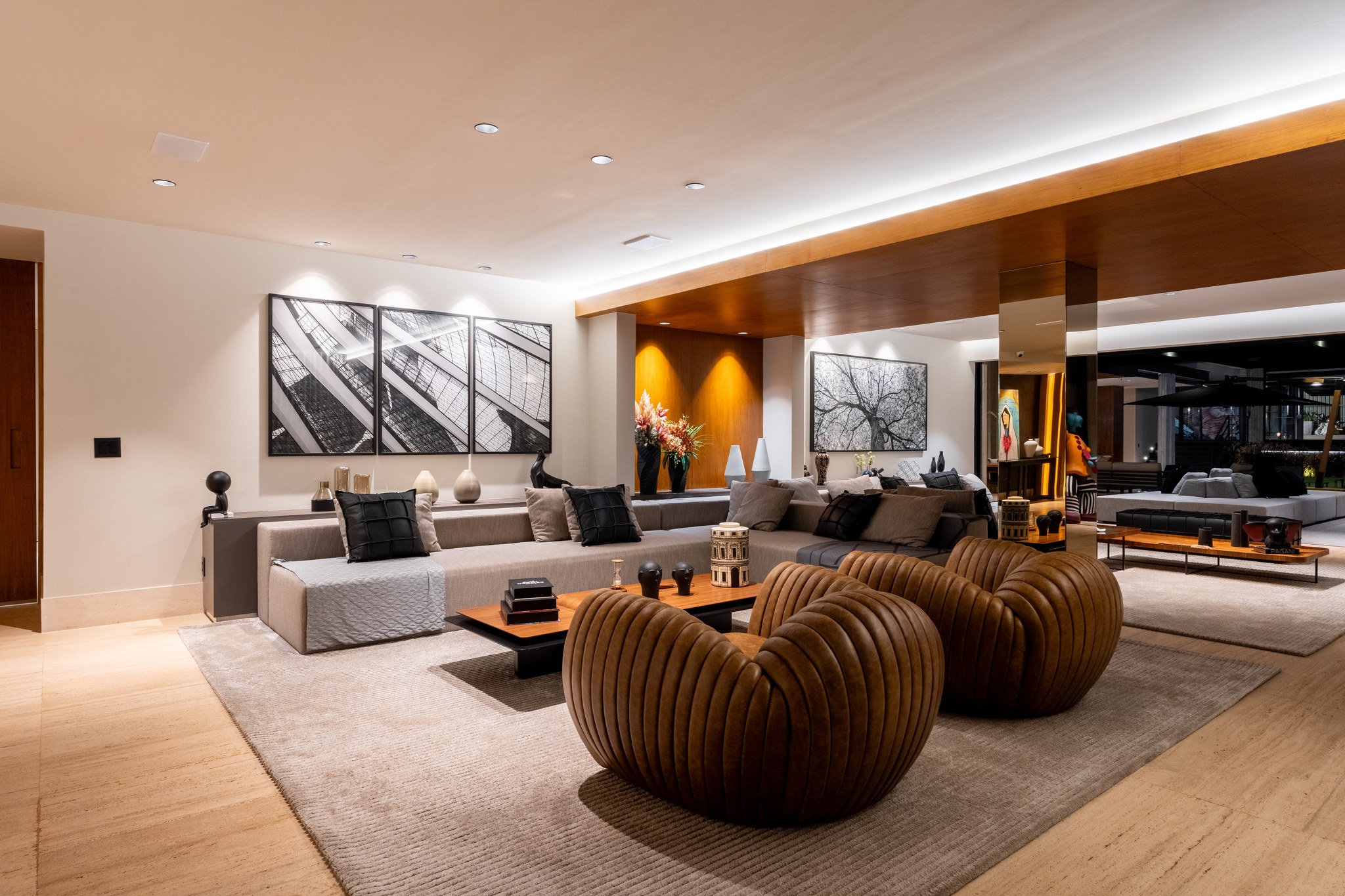 Sala de estar à noite, com poltronas de couro caramelo, sofá modular cinza e fotografia de árvore
