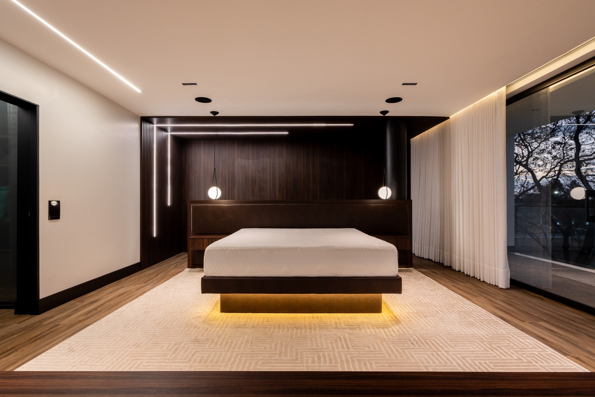 Suíte máster à noite, com cama suspensa iluminada por led, painel de madeira ripada e vista para o jardim
