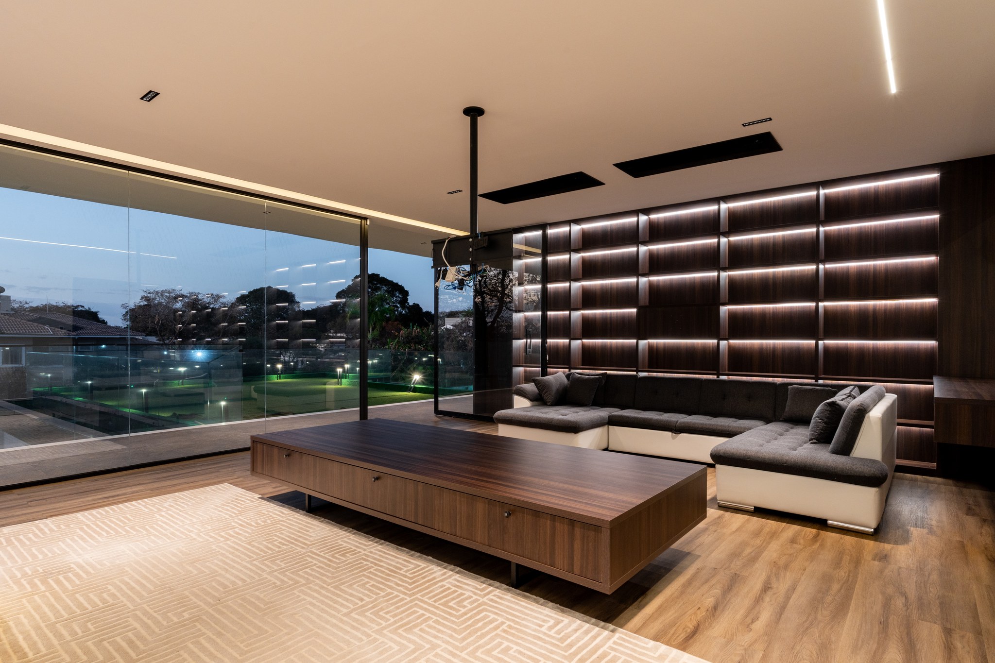 Sala de TV à noite, com sofás modulares, painel de madeira retroiluminado e vista para o campo de golfe