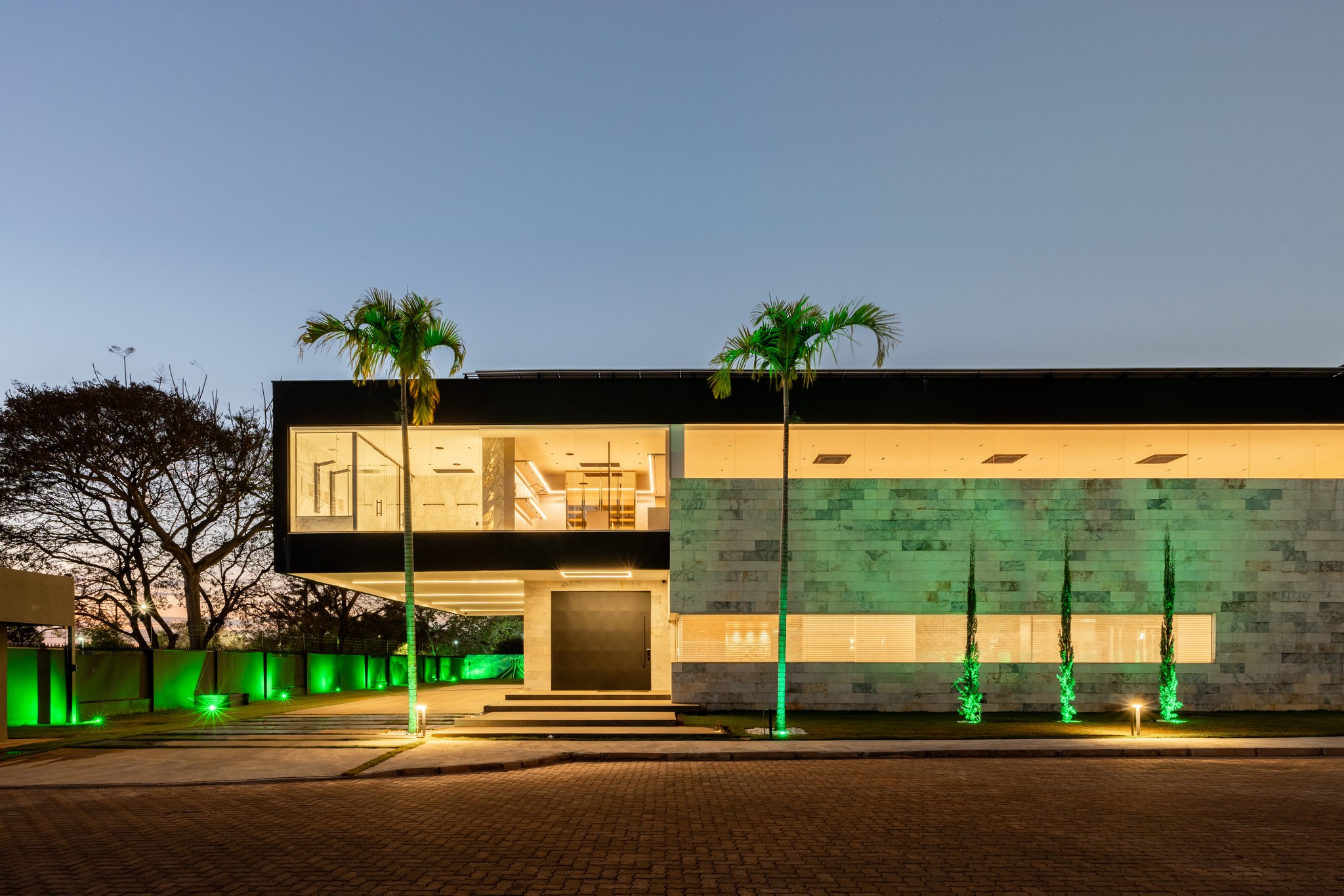 Fachada frontal ao entardecer, com revestimento em pedra verde, palmeiras imperiais e iluminação de jardim
