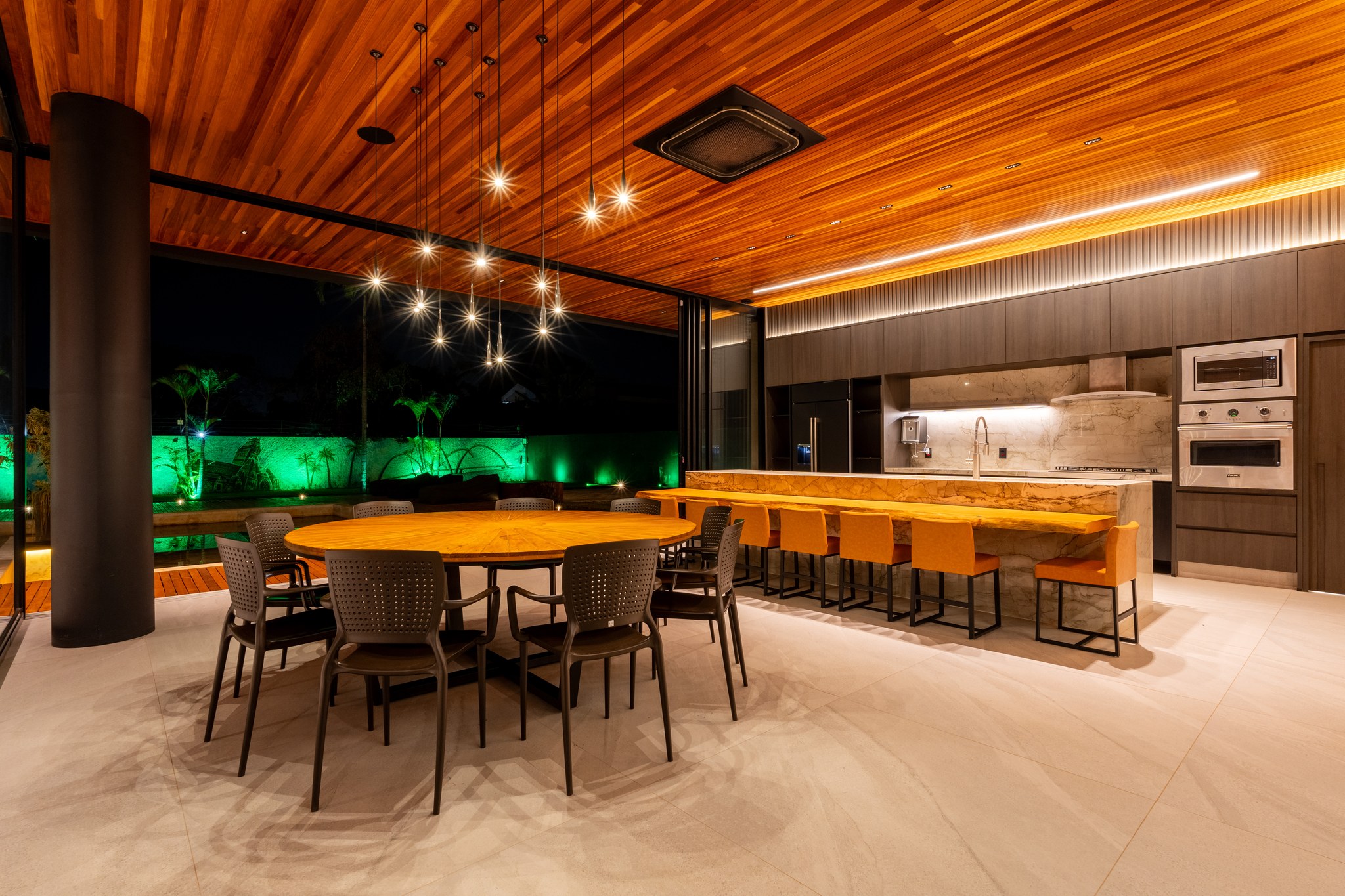 Área gourmet iluminada à noite, com mesa redonda para oito, bancada em mármore e banquetas laranja