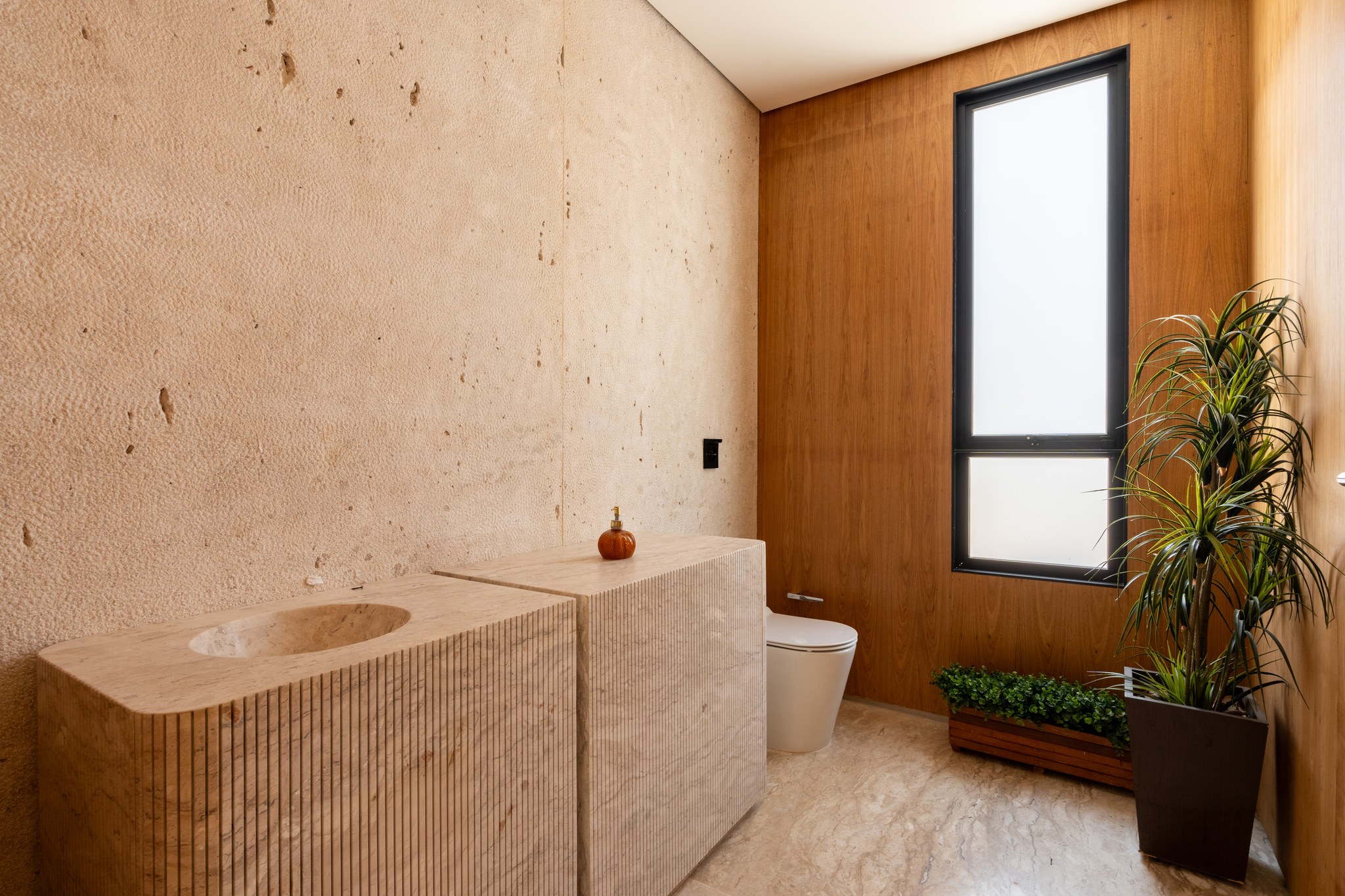 Lavabo com bancada esculpida em travertino ripado, parede em concreto aparente e planta tropical