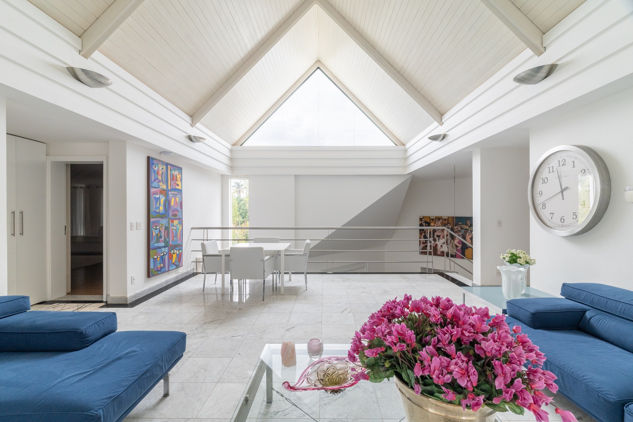 Sala de estar de pé-direito em duas águas, com claraboia central, mesa de jantar e obras de arte coloridas