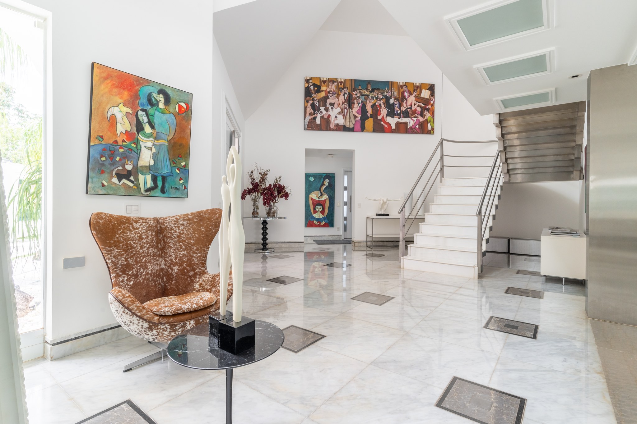 Hall de entrada com escada metálica suspensa, obras de arte figurativas e poltrona em pelego