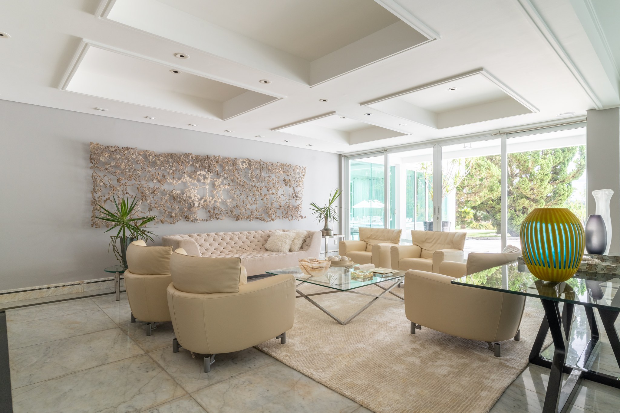 Sala de estar com poltronas em couro bege, sofá capitonê e painel escultórico em metal dourado na parede