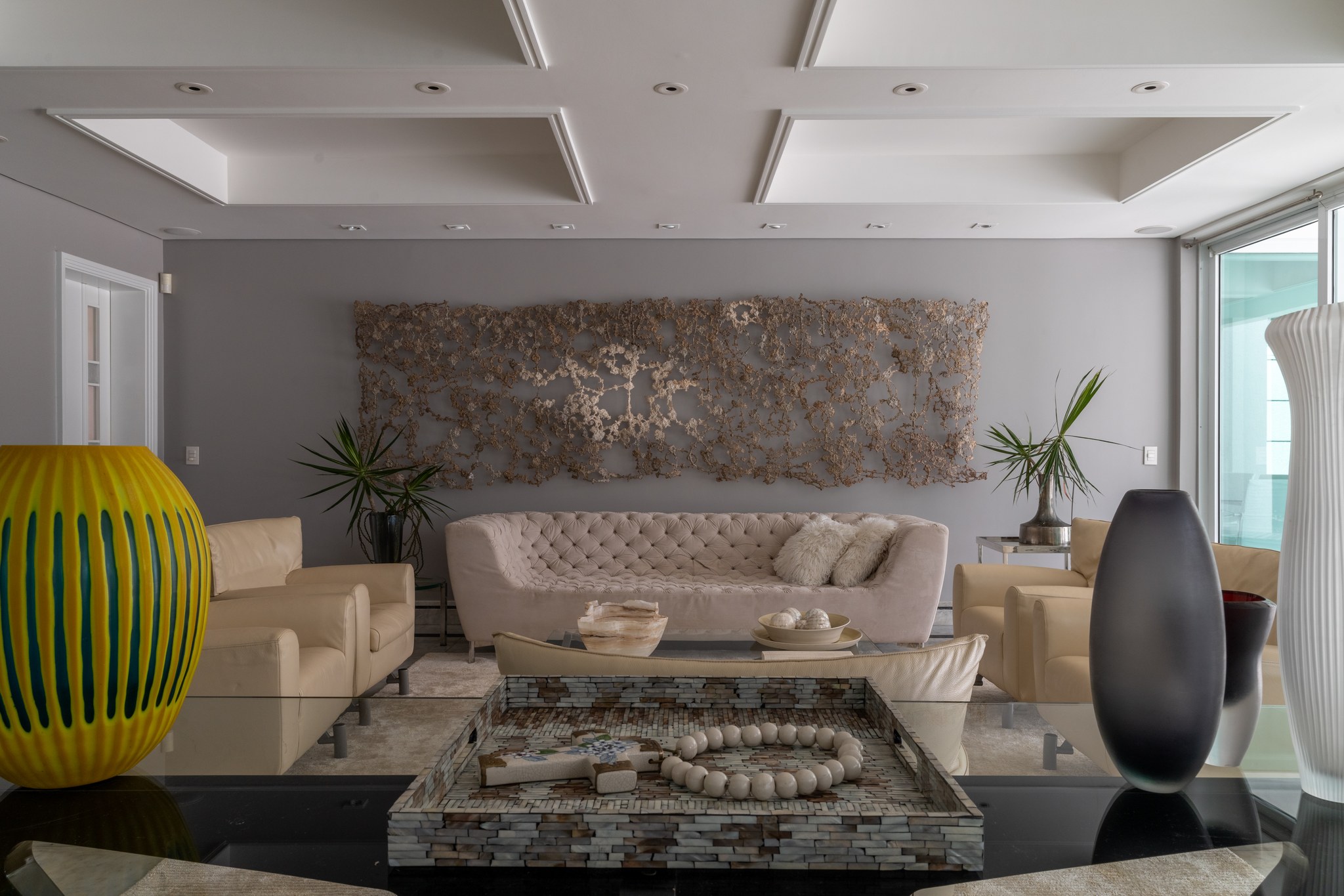 Sala de estar com vaso Murano amarelo e azul, sofá capitonê e painel escultórico dourado ao fundo